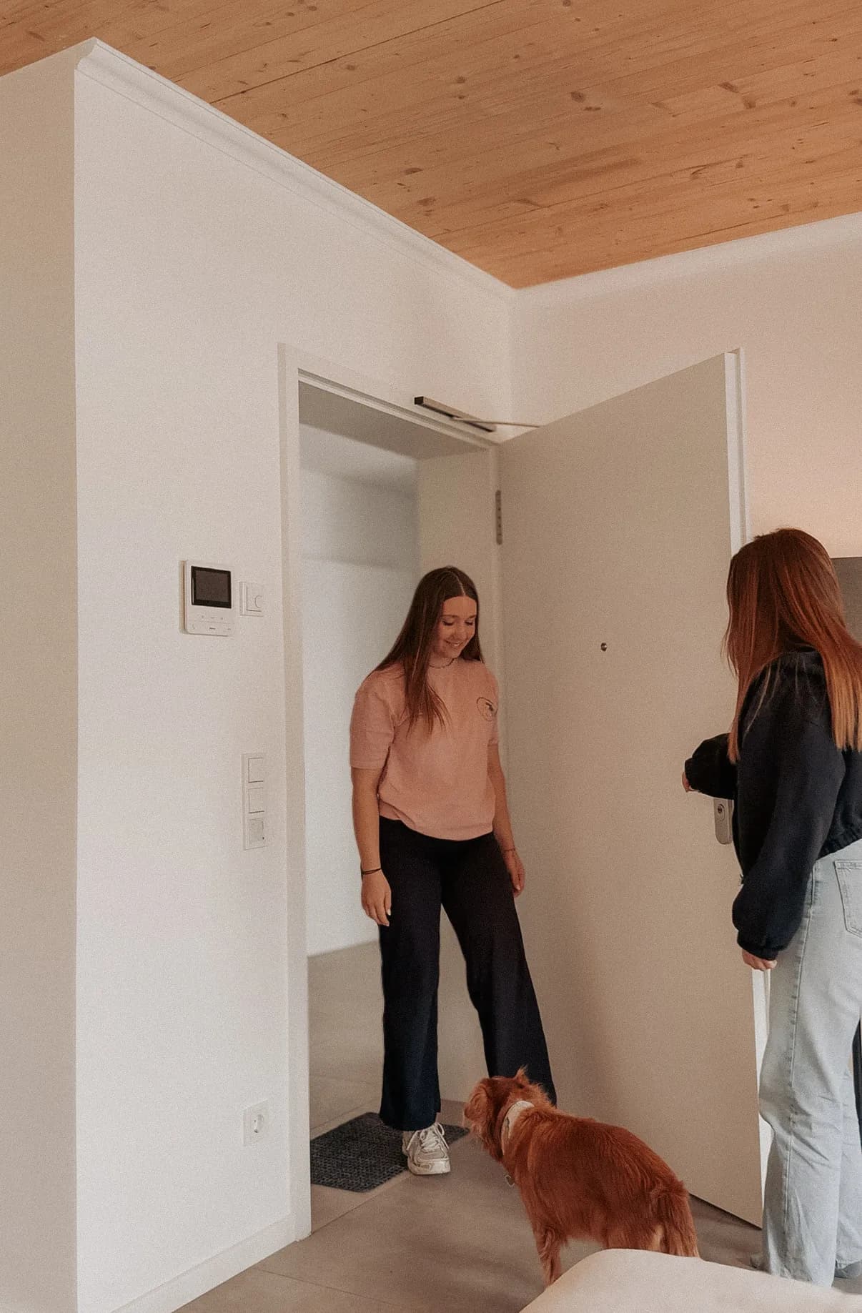 Eine Frau kommt durch die Wohnungstür herein, eine zweite hält ihr die Tür auf, ein rotbrauner Hund läuft ihr entgegen