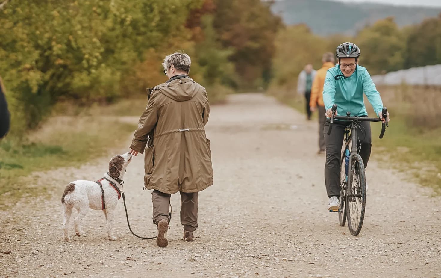Hundehalterin geht mit ihrem angeleinten Hund auf dem Feldweg an einer entgegenkommenden Radfahrerin vorbei